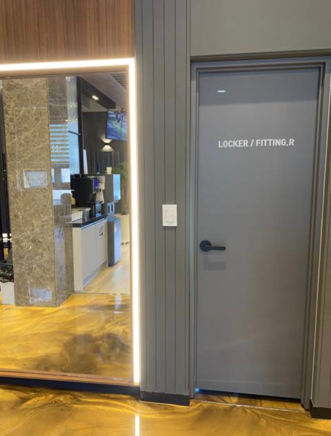 복도에서 본 라운지. 거울 너머로 커피 머신과 정수기가 보이고 오른쪽은 LOCKER / FITTING.R 문입니다.