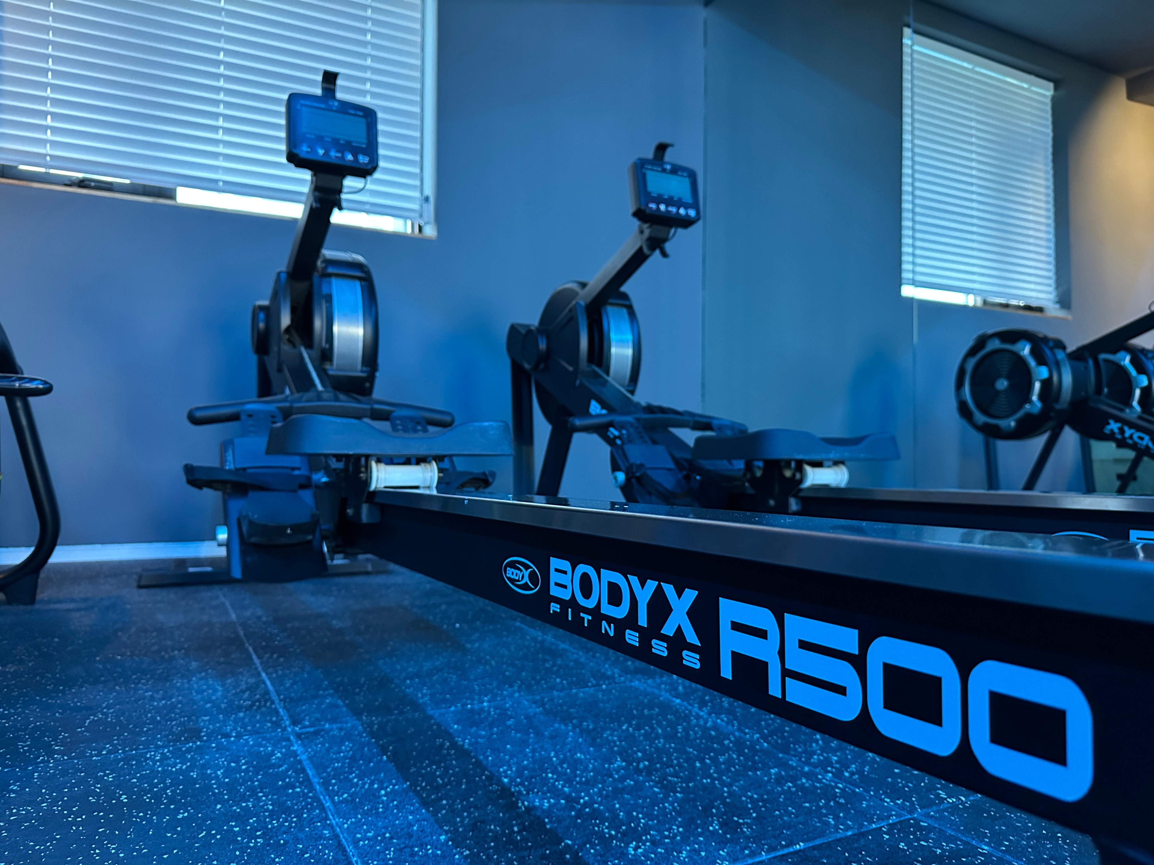 로잉머신 열. BODYX R500 로잉머신이 나란히 놓여 있고 창가로 블라인드가 내려와 있습니다.
