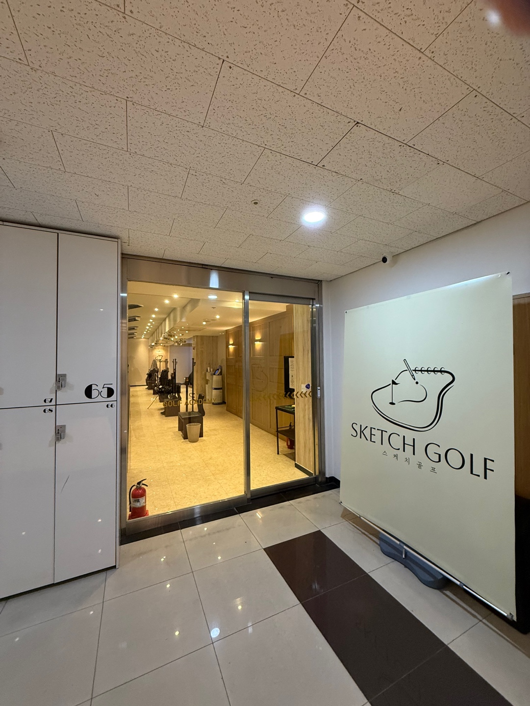 상가 지하 복도에서 본 매장 입구. 유리 자동문 옆에 SKETCH GOLF 배너가 서 있습니다.